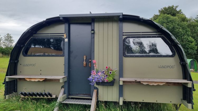 O mică casă mobilă, denumită Landpod, cu un design curbat, situată pe un teren verde, având o ușă centrală și feronerie decorativă, cu flori colorate pe terasa din față.