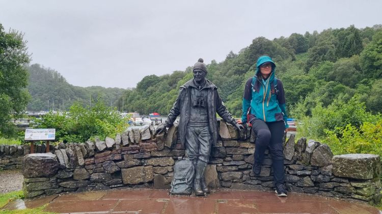 O femeie îmbrăcată într-o jachetă waterproof stă lângă o statuie din bronz a unui bărbat în echipament de pescuit, pe un zid de piatră, în fața unui peisaj natural verde, în condiții de vreme ploioasă.