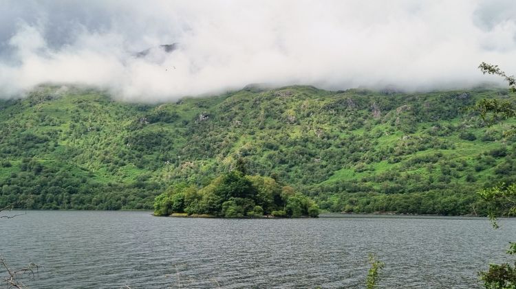Peisaj cu un lac liniștit, înconjurat de coline verzi acoperite de nori.